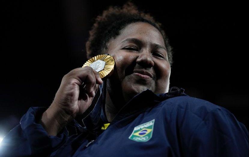 Betriz Souza é uma mulher negra e alta de cabelos crespos amarrados em um coque. Na foto, ela sorri e exibe sua medalha de ouro conquistada nas Olimpíadas de Paris