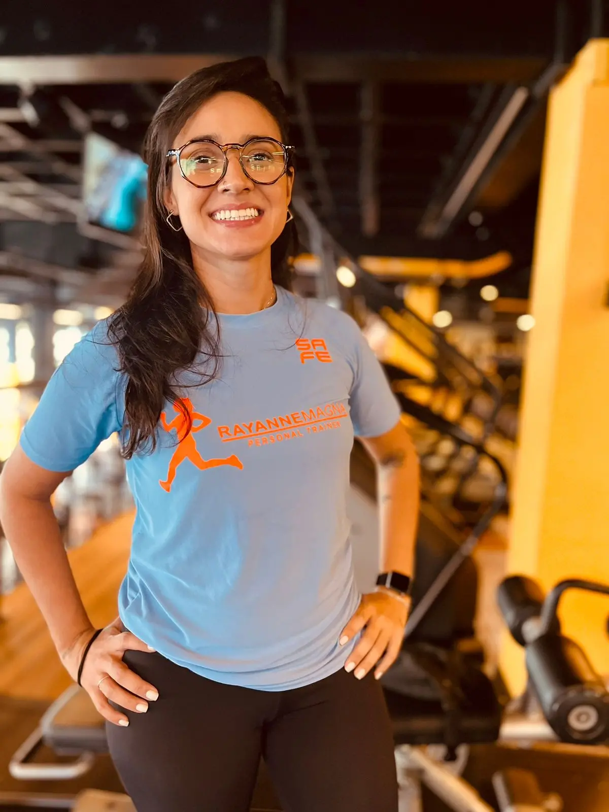 Rayanne Magna é uma mulher magra de longos cabelos pretos. Ela usa óculos redondos de armação preta e, na foto, está com camisa azul e calça preta