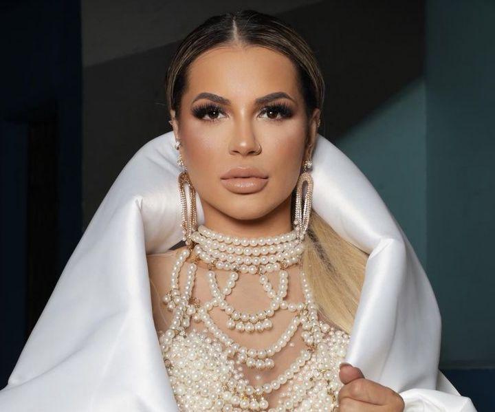 Deolane usando uma roupa branca com pérolas. Ela é uma mulher loira com cabelo longo e liso