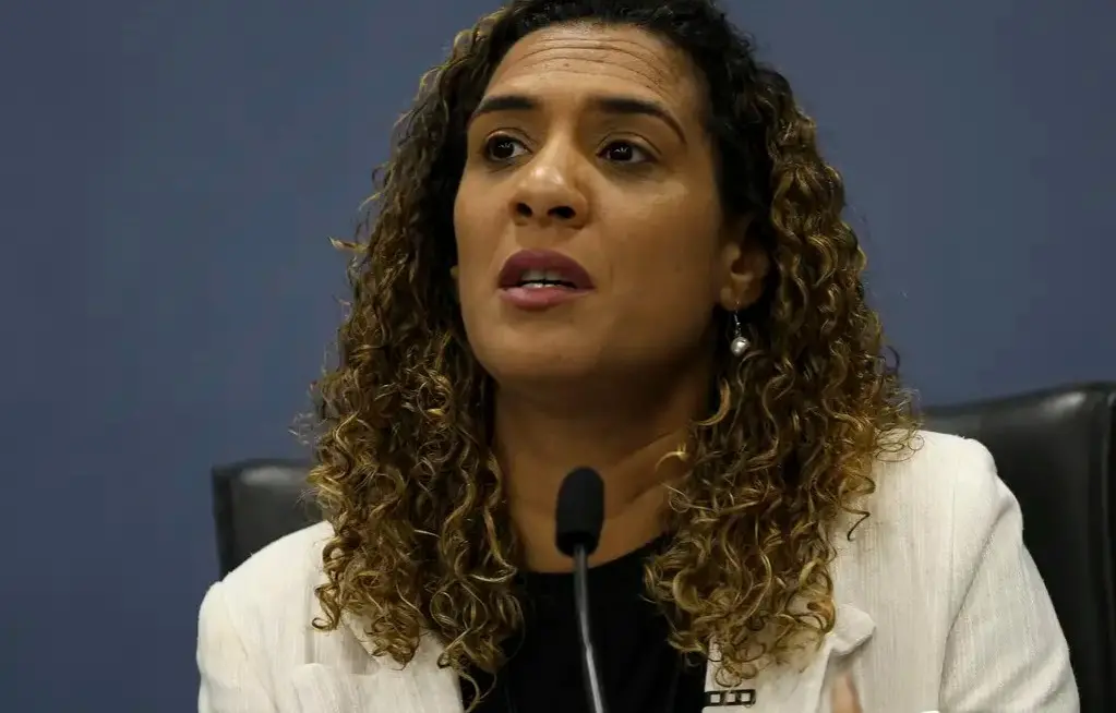 Anielle Franco é uma mulher negra de cabelos cacheados iluminados. Na foto, ela está de terno branco e camisa preta, discursando