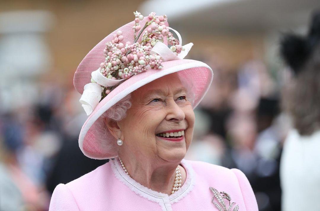 Imagem mostra a rainha Elizabeth II sorrindo. Ela está usando chapéu rosa e uma roupa rosa.