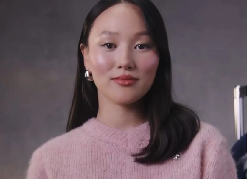 Yerin Ha é uma atriz australiana. Ela é uma mulher branca, de cabelos longos, pretos, e está usando uma maquiagem em tons de rosa. Na foto, ela também está usando um moletom rosa