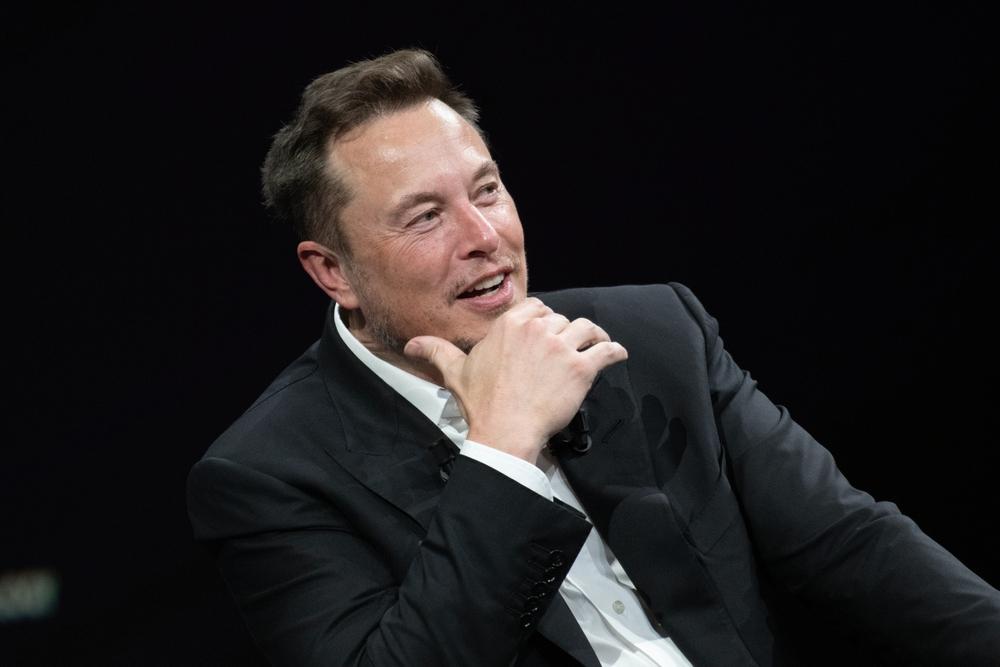 Elon Musk é um homem branco de meia idade e cabelo preto curto. Na foto, ele está pensativo, discursando, de terno e camisa branca por baixo