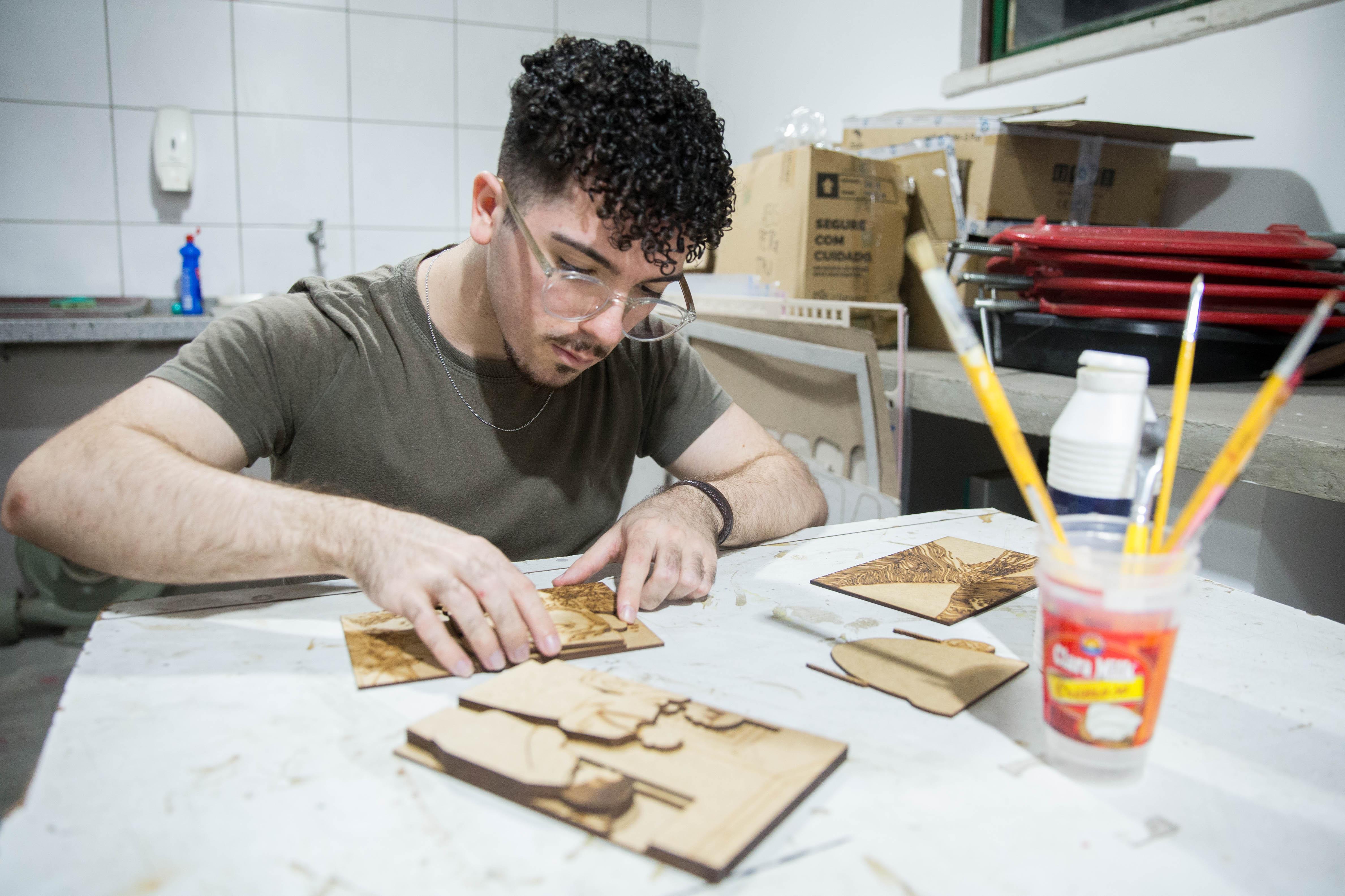 Homem colando diferentes partes de uma peça tátil, e uma mesa com materiais artísticos, como pinceis