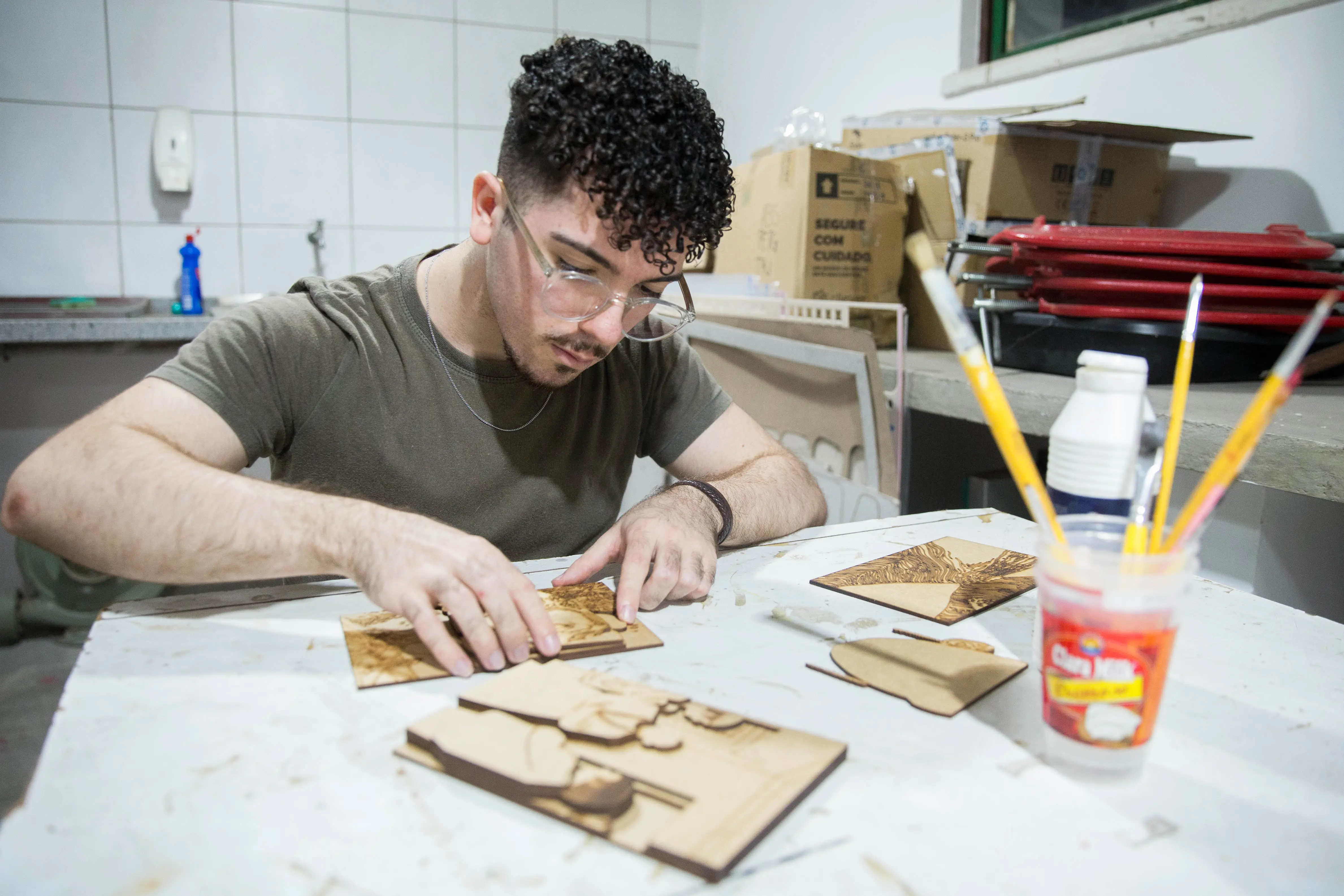 Homem colando diferentes partes de uma peça tátil, e uma mesa com materiais artísticos, como pinceis