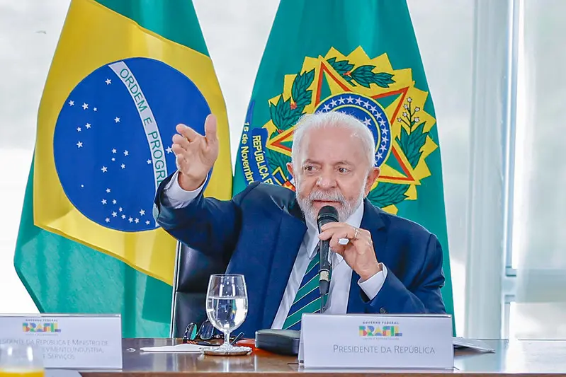 Lula é um homem branco, idoso, de cabelos também brancos. Na foto, ele está discursando enquanto segura um microfone. Ao fundo, está a bandeira do Brasil