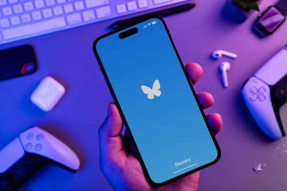 Homem segurando um smartphone iPhone 14 Pro com aplicativo de mídia social Bluesky com controladores de jogos de vídeo PS5. Vista superior