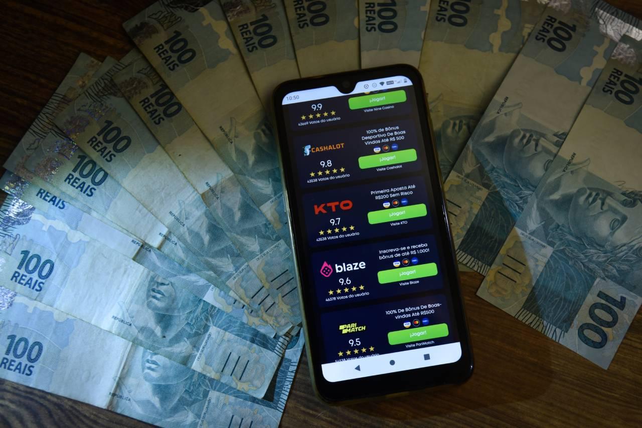 Celular mostrando aplicativo de bet sobre várias notas de R$ 100