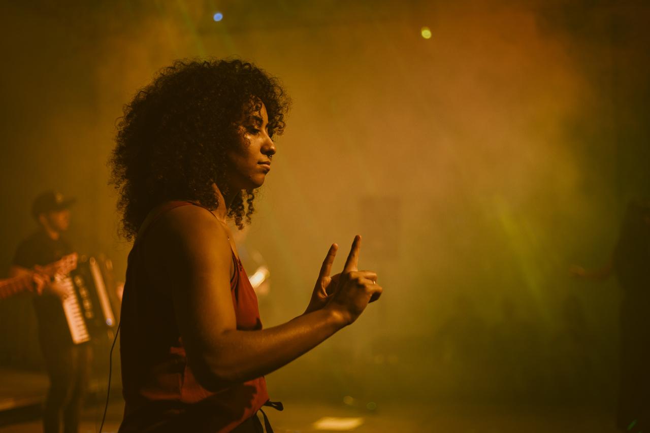 Uma mulher jovem, de pele negra e cabelos cacheados pretos, interpreta sinais em um palco. O ambiente está iluminado por uma luz amarela