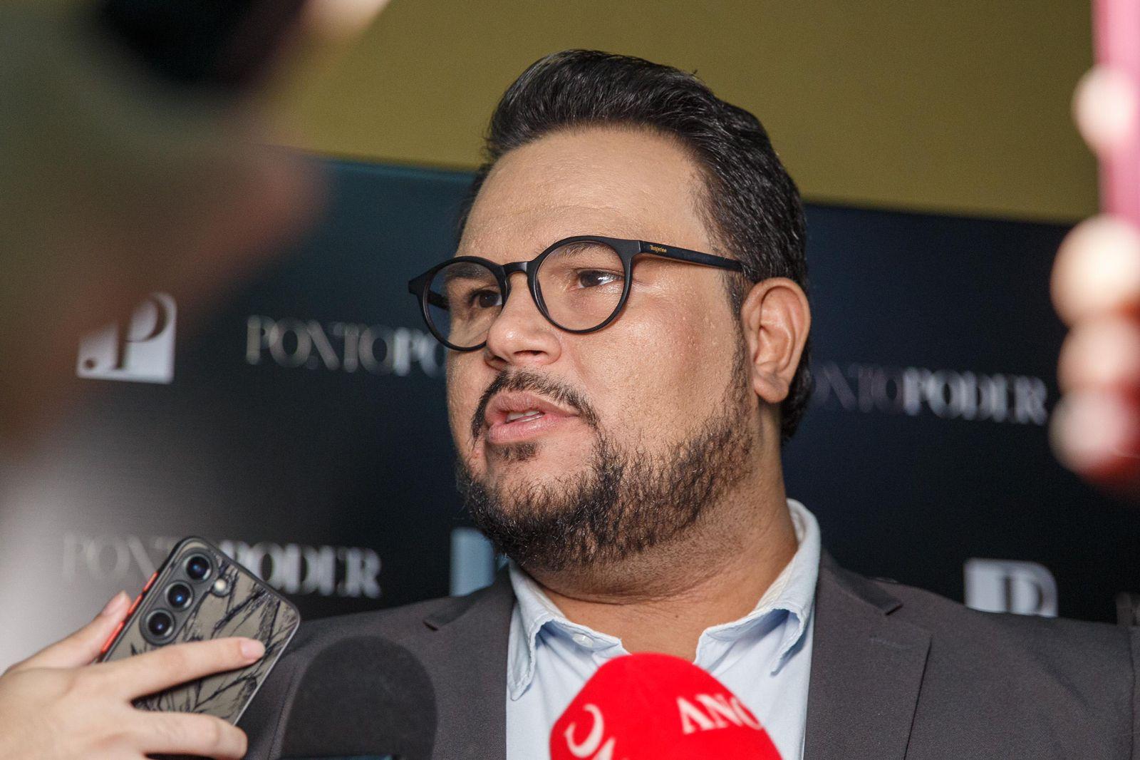 Técio Nunes é um homem alto, de barba bem feita e cabelo curto. Na foto, ele está de óculos de grau de armação preta e usa terno cinza e camisa azul de botão