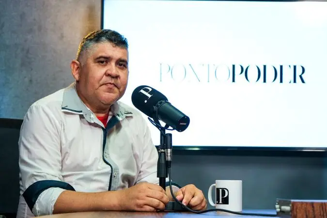 Homem de cabelos pretos com blusa social clara, sendo entrevistado.