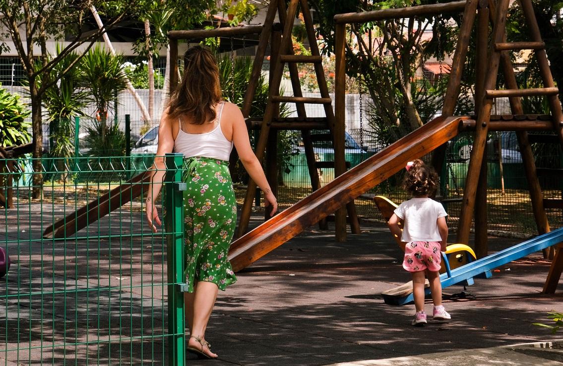 Mãe anda atrás de filha criança em parque de madeira de praça pública