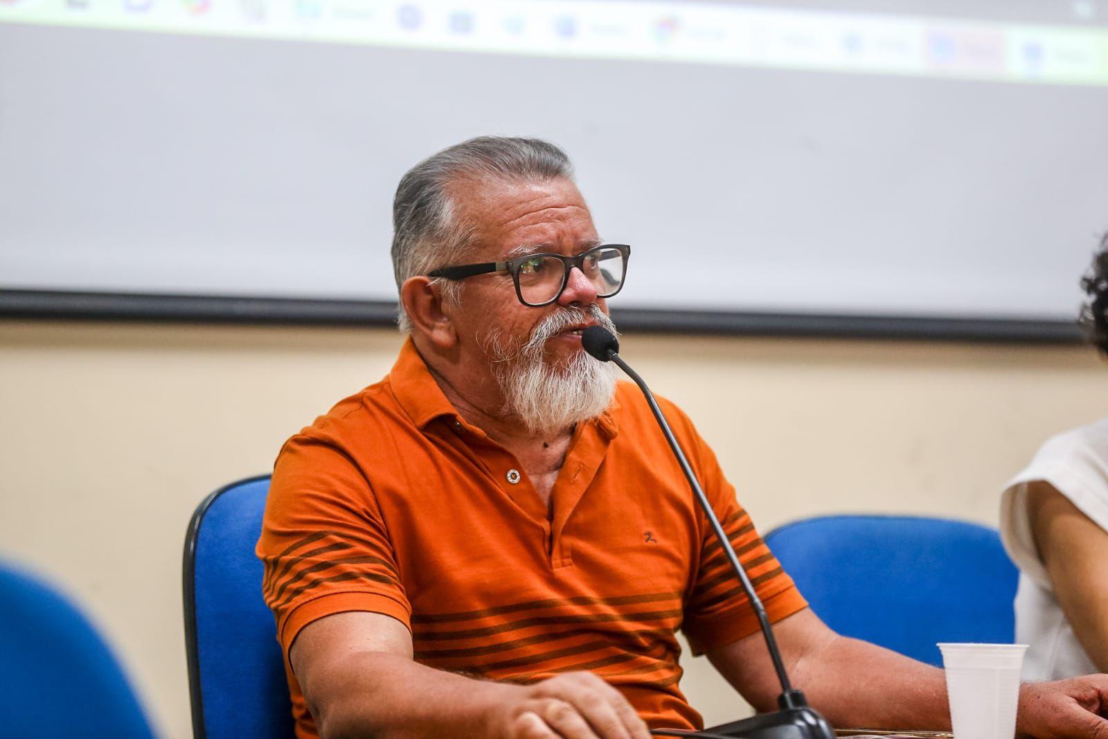 Chico Malta é um homem branco, idoso e de camisa polo laranja. Ele também usa óculos de armação preta