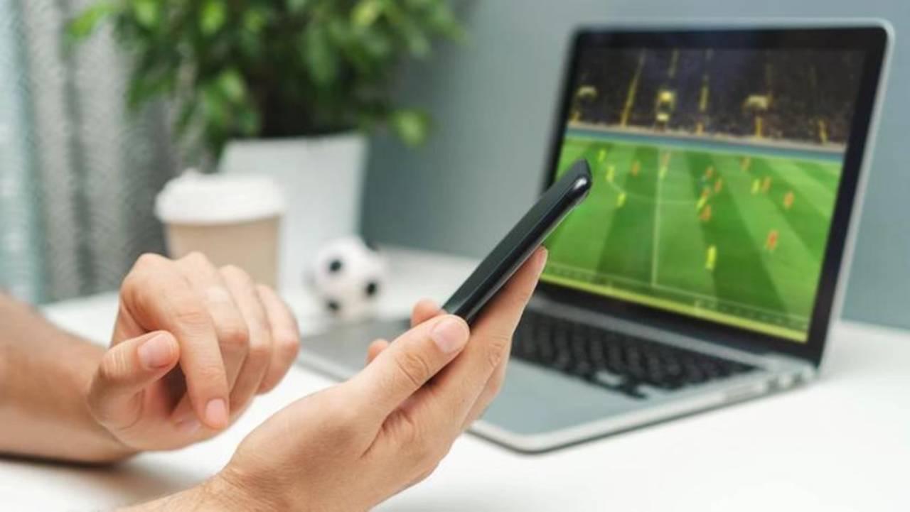 Mãos com celular e computador ao fundo mostrando partida de futebol