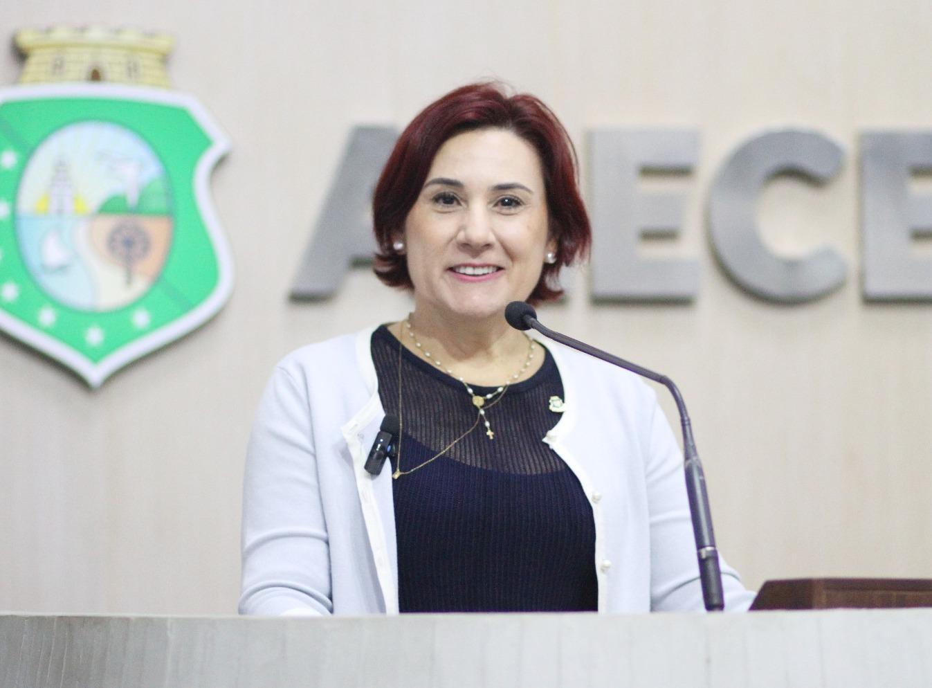 Emília é uma mulher branca, idosa, de cabelo curto liso e ruivo. Na foto, ela está de blusa preta por baixo de um blazer branco em auditório da Assembleia Legislativa