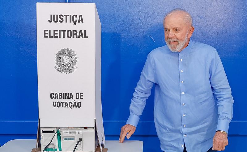 Lula é um homem branco, idoso e de cabelo branco e barba branca. Na foto, ele está de camisa azul de botão saindo de uma cabine de votação