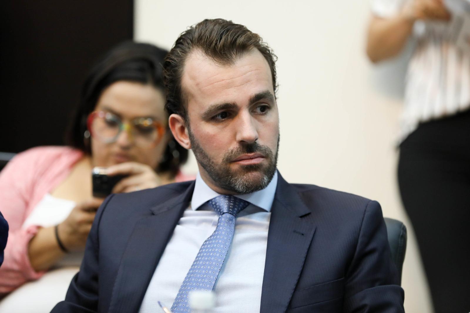 Lucas Bove é um homem branco, calvo, de cabelo penteado para trás. Na foto, ele está d eterno preto, camisa de botão azul e gravata azul