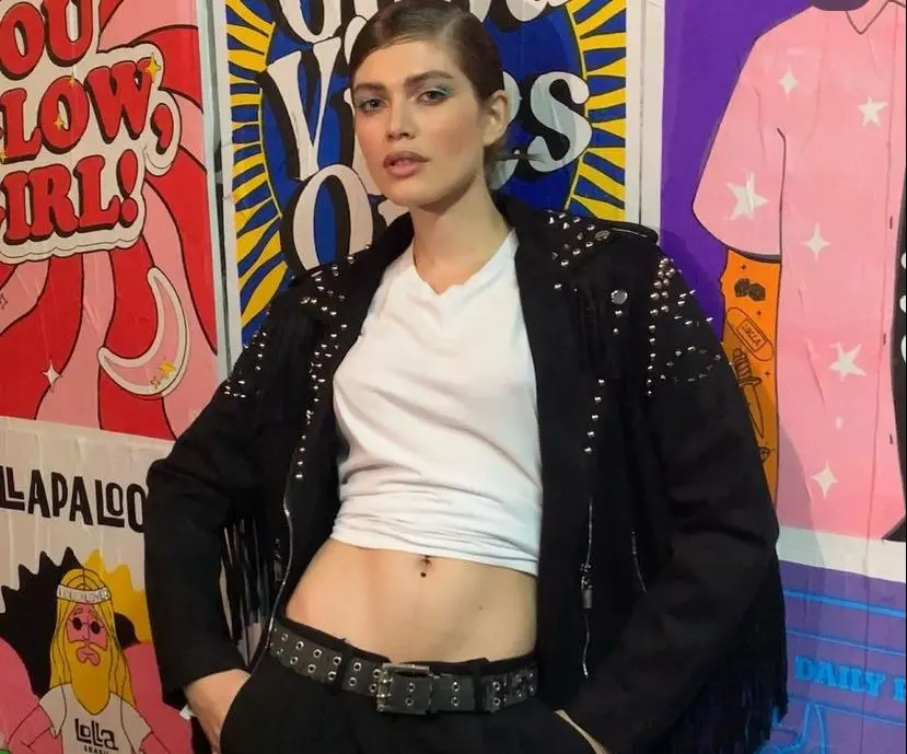 Valentina é uma mulher transgênero branca, magra, e de cabelos loiros. Na foto, ela está com o cabelo amarrado e usa uma blusa branca, calça jeans com cinto largo e jaqueta preta