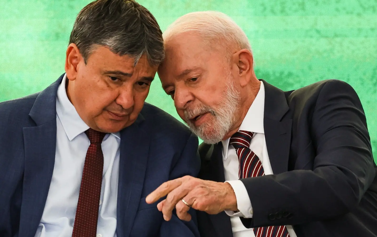Wellington Dias e Lula estão sentados um ao lado do outro, conversando. Lula fala enquanto Wellington ouve. Os dois estão de terno e camisa social