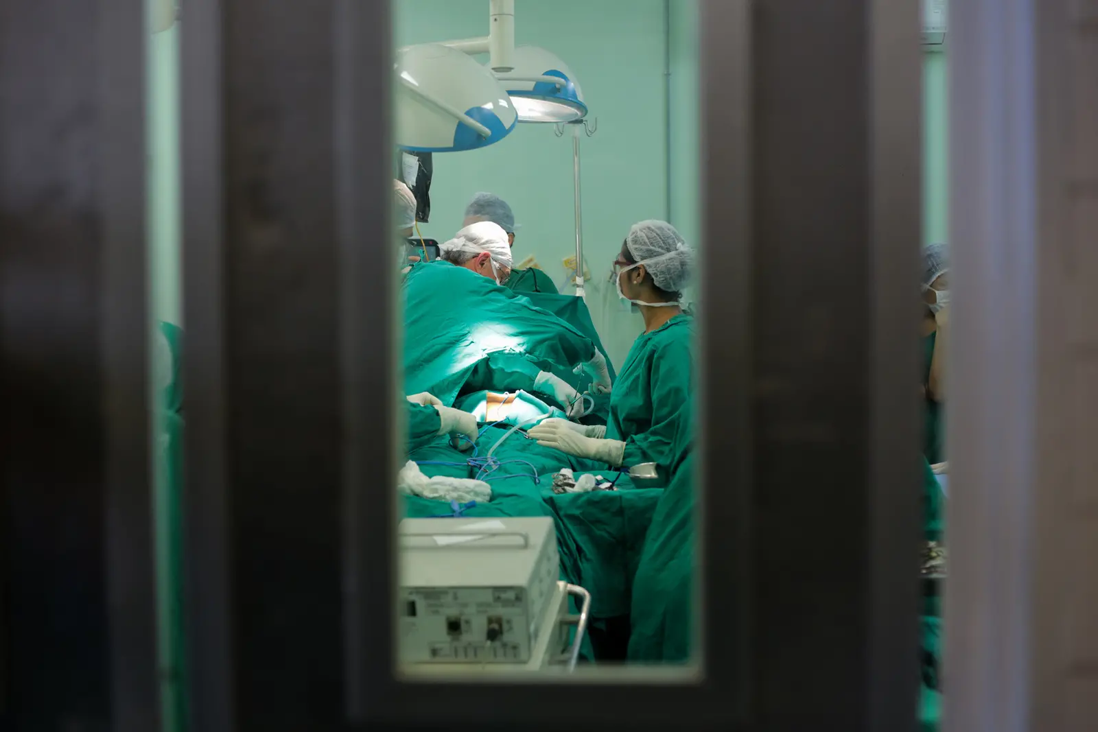 Visão de cirurgia de transplante com médicos paramentados, por meio de uma janela de vidro na porta da sala