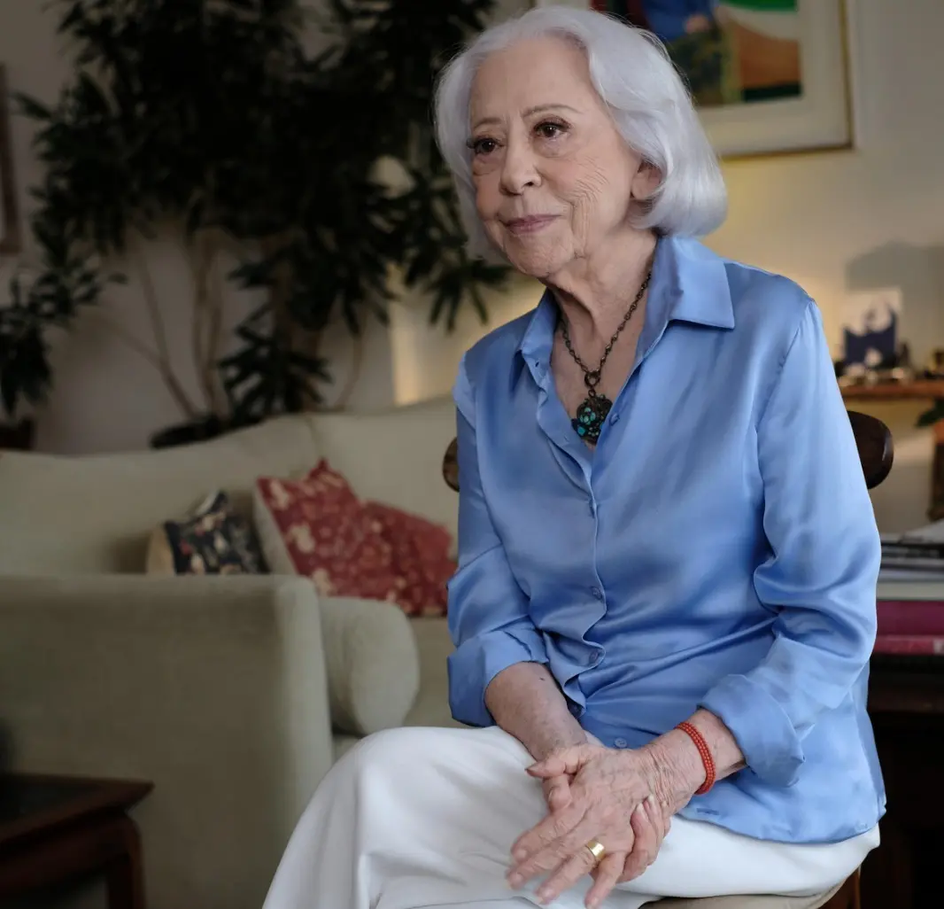 Fernanda Montenegro é uma mulher idosa branca e de cabelos também brancos cortados em chanel. Na foto, ela está de camisa de botão azul e calça branca