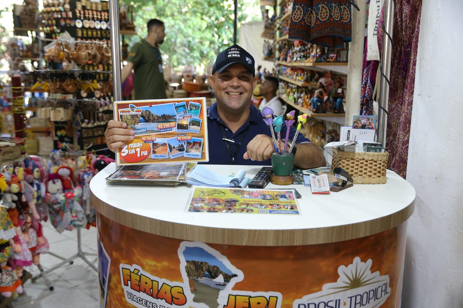 Diógenes posa no stand onde vende pacotes de viagem. Ele segura um mostruário com valores
