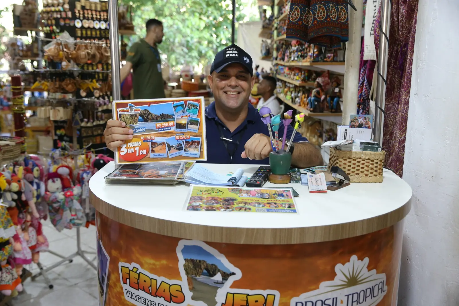 Diógenes posa no stand onde vende pacotes de viagem. Ele segura um mostruário com valores