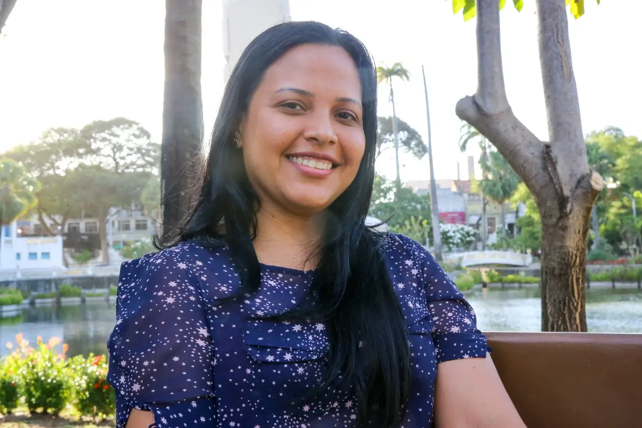 Eimily, mulher venezuelana de cabelos pretos e lisos, sorrindo para a câmera. Ao fundo, paisagem de parque de Fortaleza
