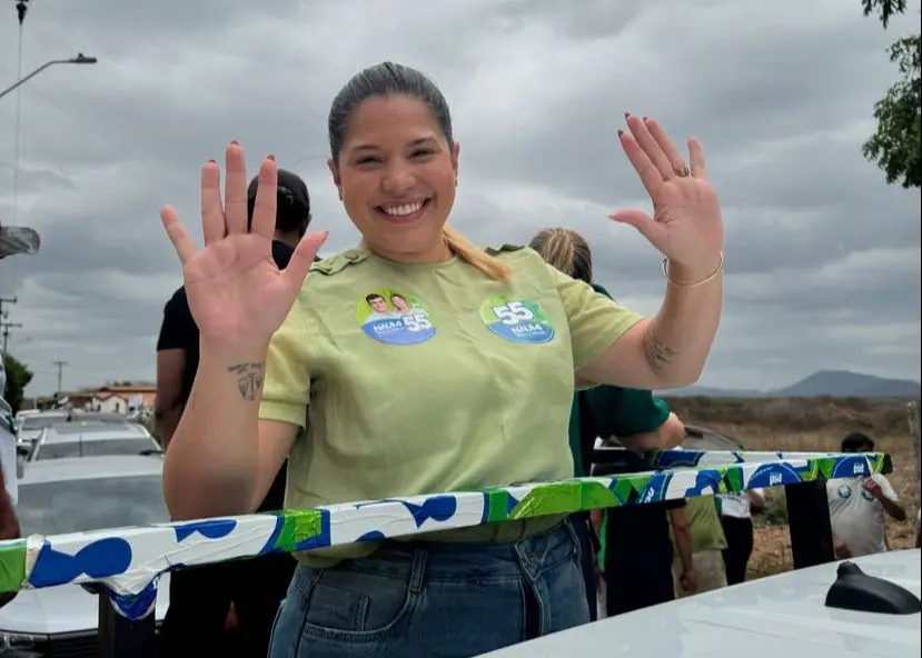 Priscila é uma mulher branca e de cabelo liso e loiro. Na foto, ela está de camiseta verde, sorrindo, fazendo 10 com os dedos