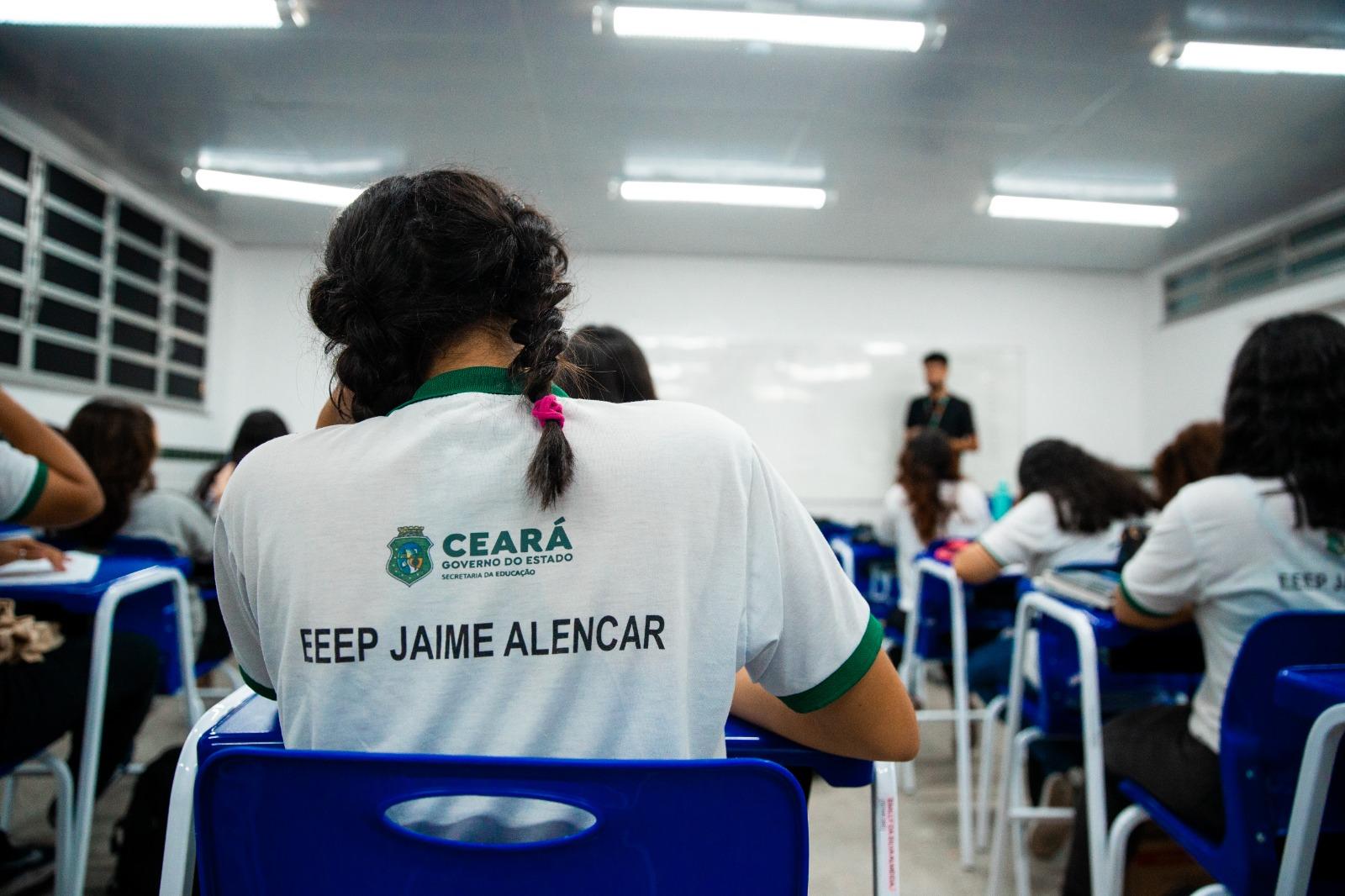 Estudante de costas, com farda em que está escrito 'EEEP JAIME ALENCAR', com a logo do governo do estado. Ao fundo, desfocado, estão outros alunos e o professor