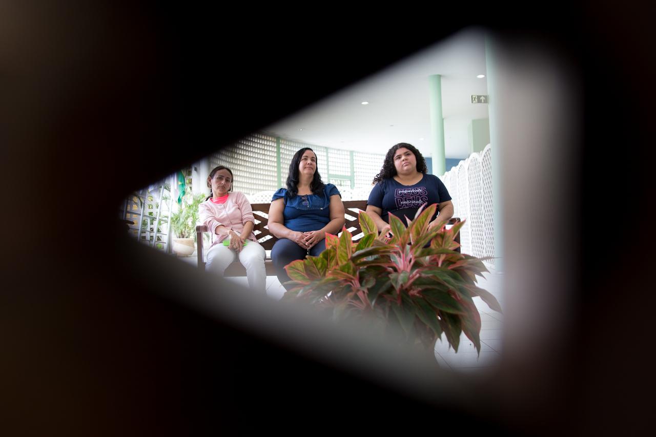 Três mulheres sentadas, acompanhando o encontro. A foto foi feita por trás de uma espécie de cobogó, em forma de triângulo, que vira uma moldura no entorno das mulheres