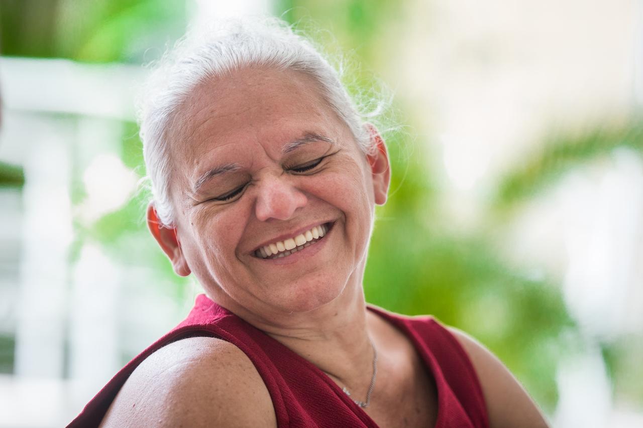 Nilde, uma mulher de cabelos brancos e usando vestido vermelho, sorri com olhos fechados