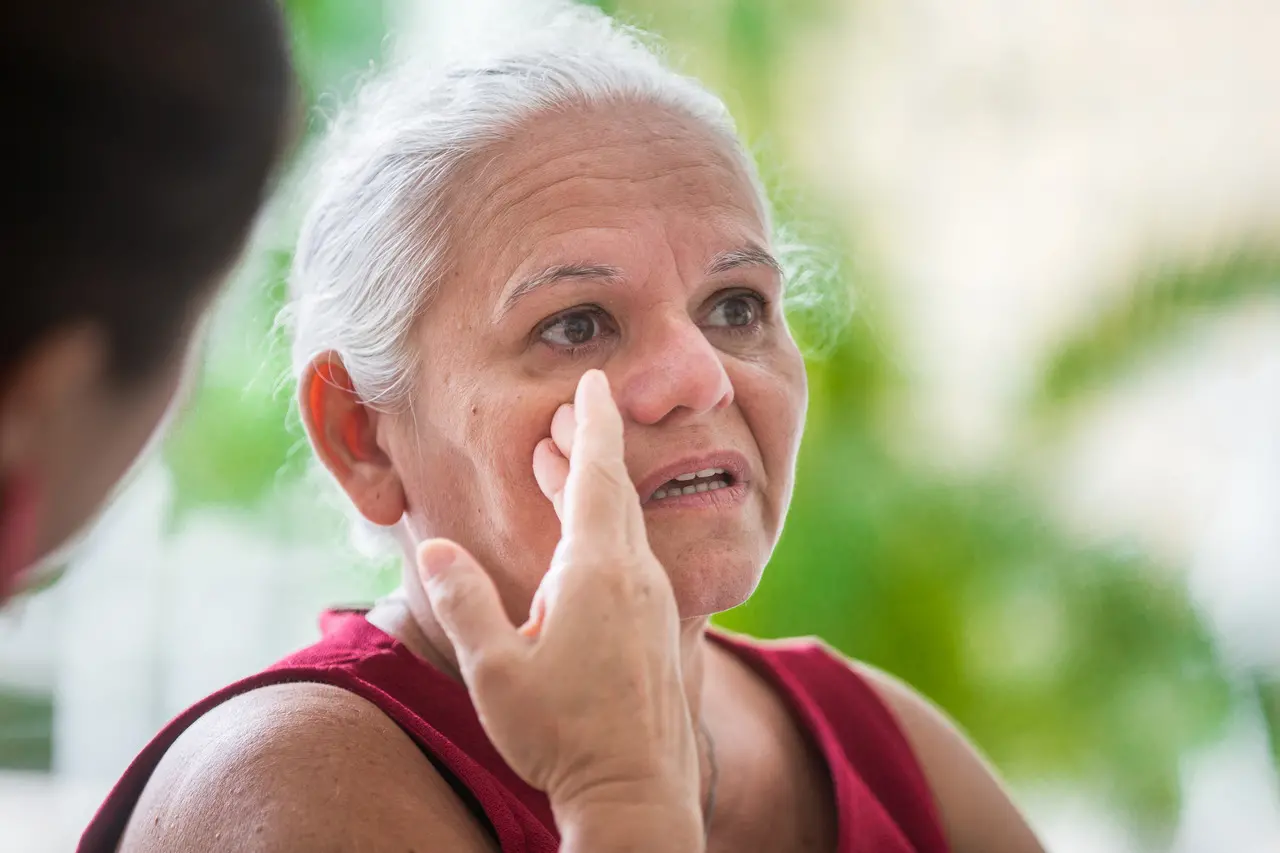 Nilde, uma mulher de cabelos brancos e usando vestido vermelho, olha para cima e limpando a lágrima do rosto