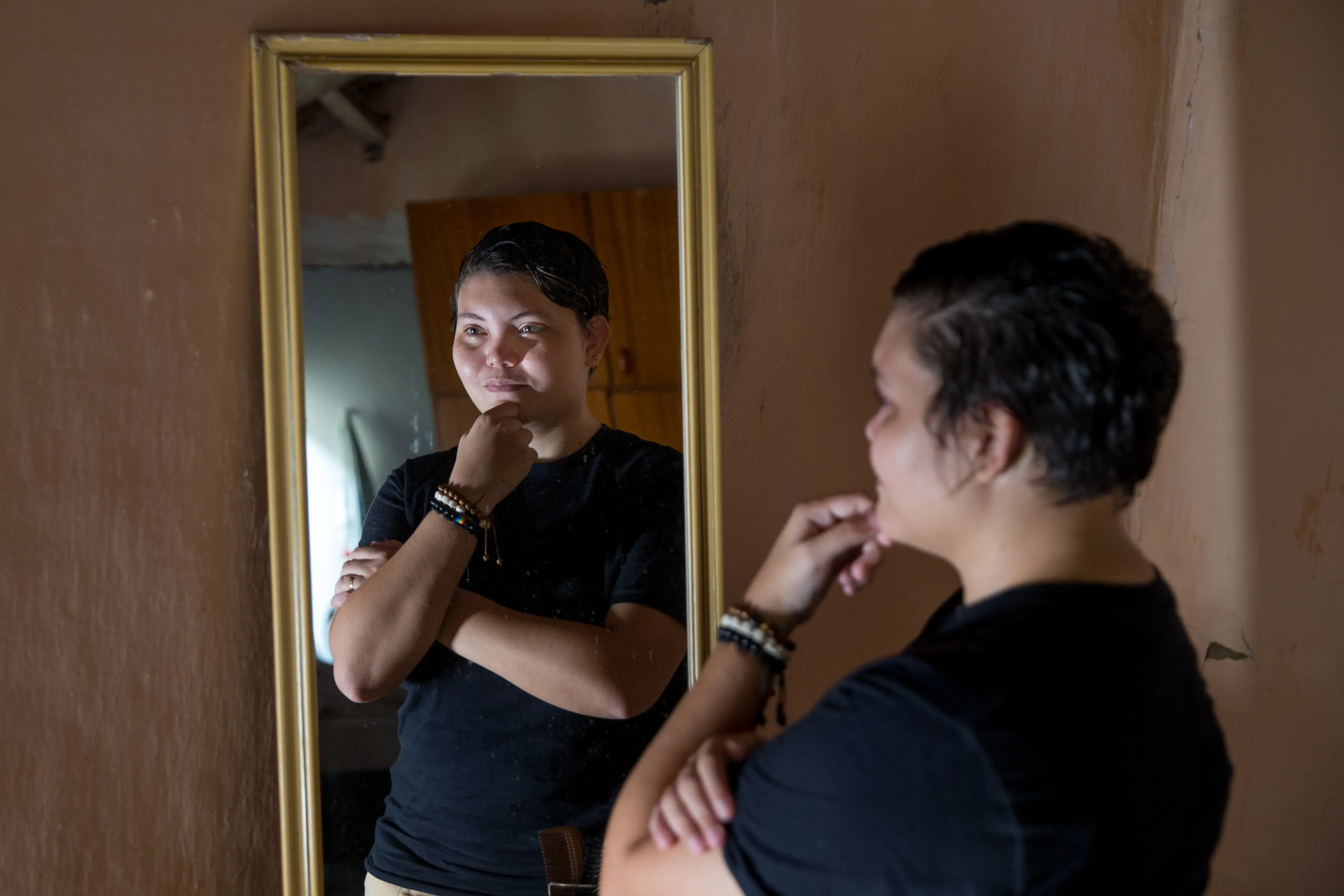 Brena se olha no espelho, com os braços cruzados e apoiando o queixo com uma das mãos