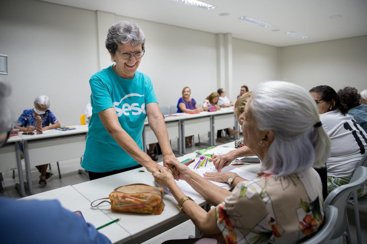 Luciene, em pé, sorri e segura as mãos de outra mulher, que está sentada