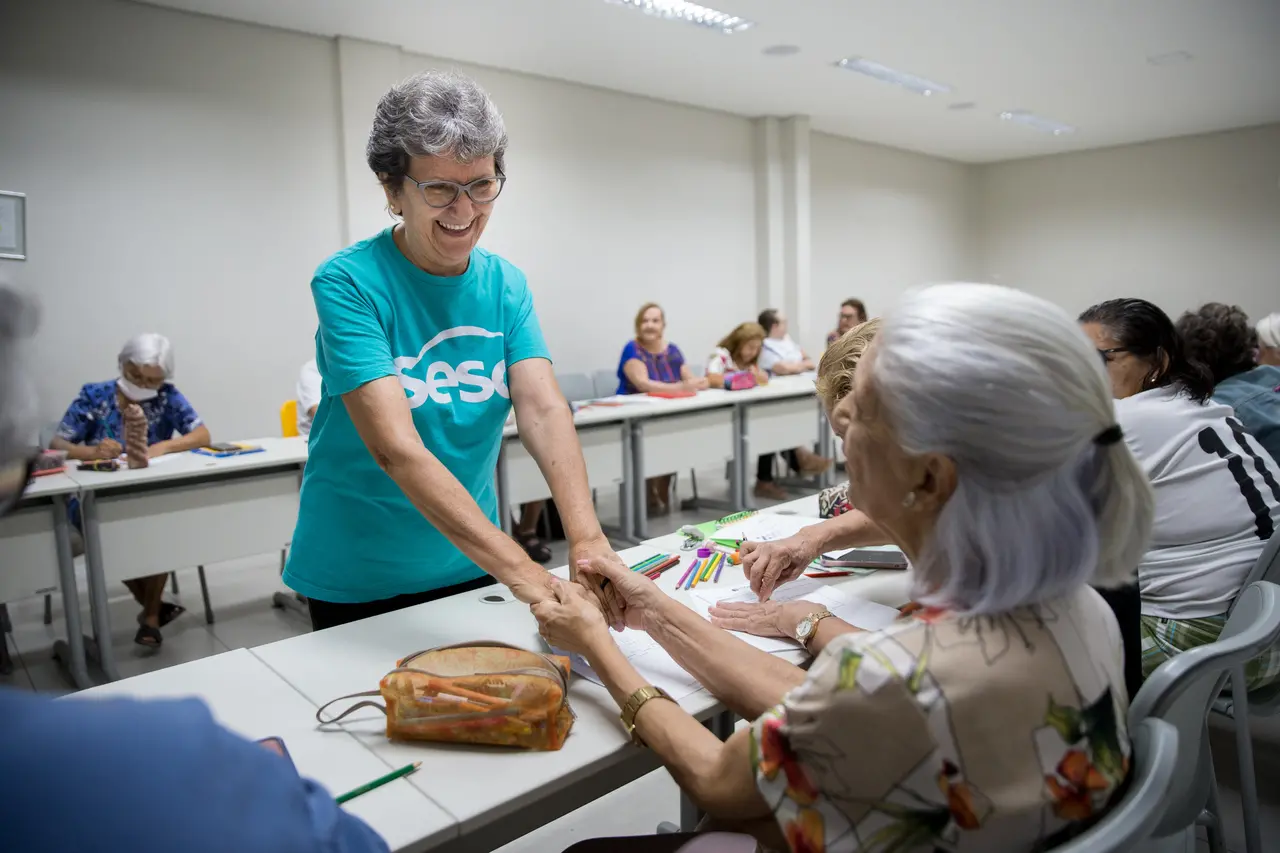 Luciene, em pé, sorri e segura as mãos de outra mulher, que está sentada
