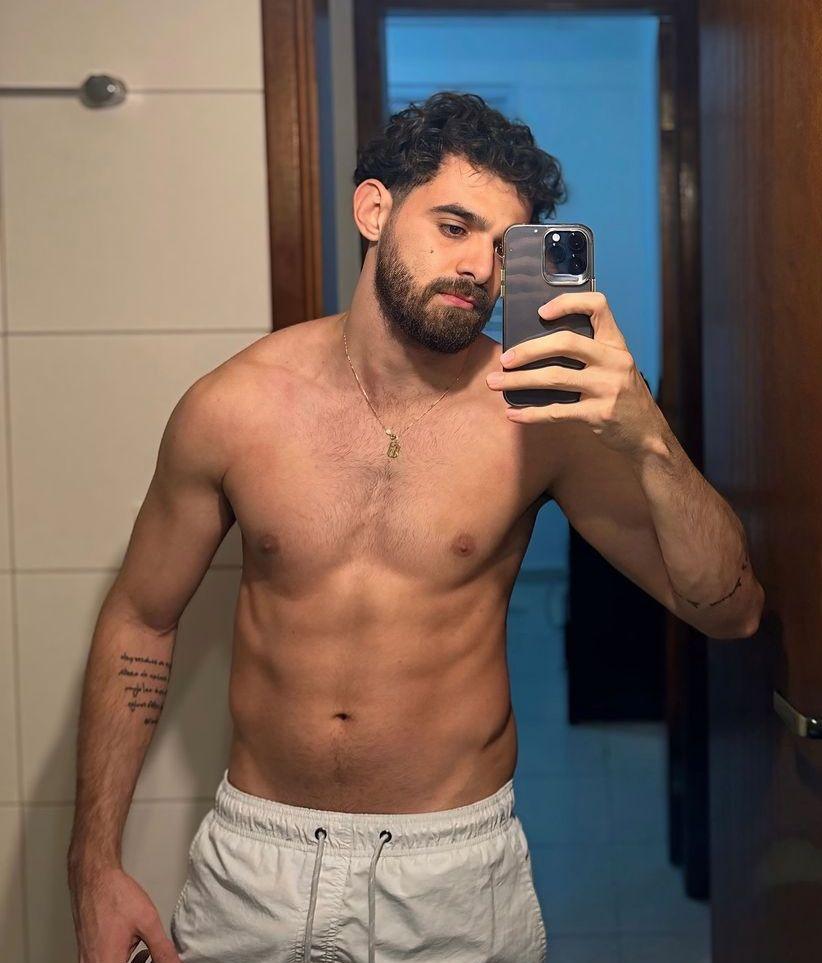 Heitor é um homem branco e bem definido. Na foto, uma selfie no espelho, ele está sem camisa, de bermuda branca e usa barba e cordão no pescoço