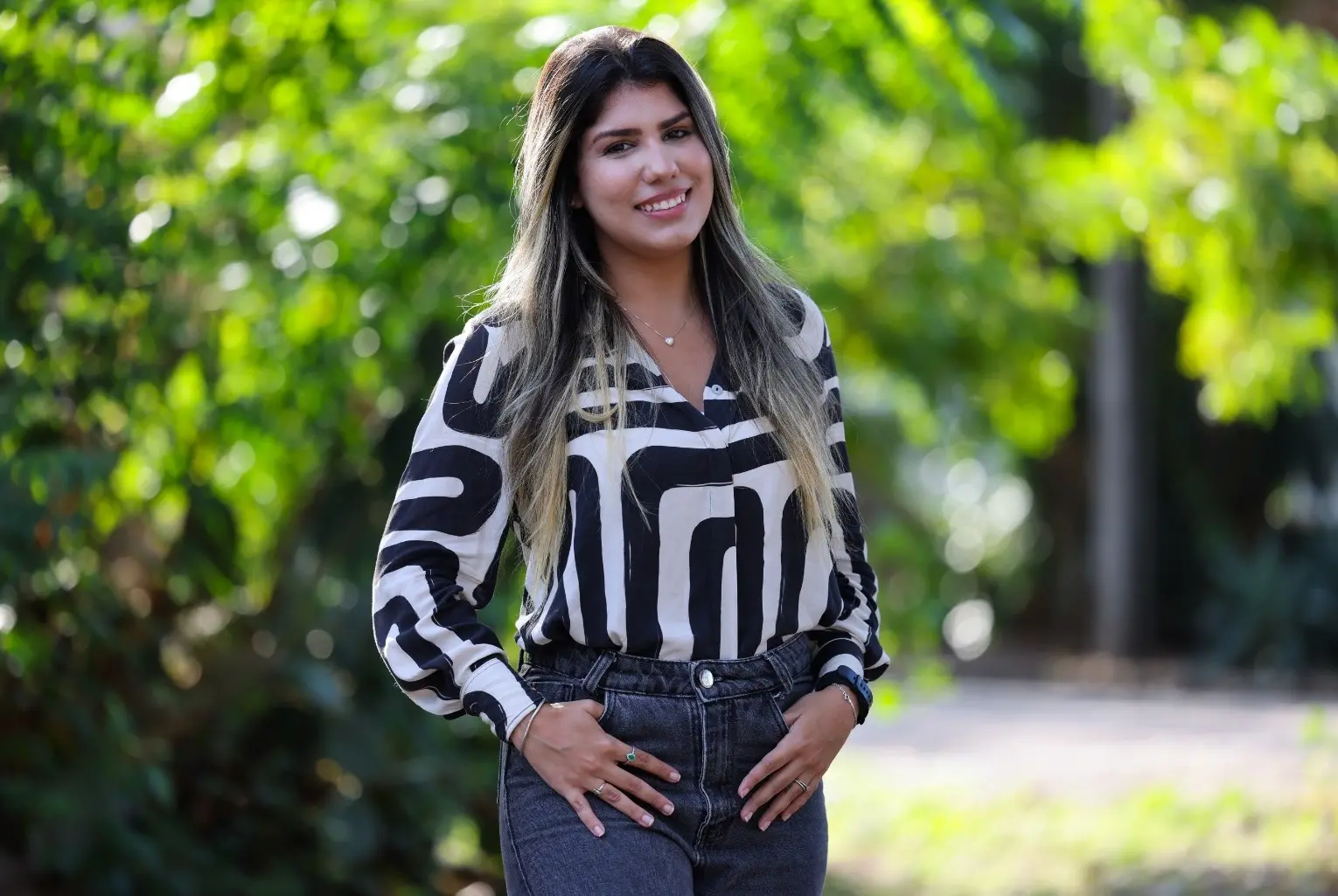 Rafaella é uma mulher branca e loira de cabelo longo. Na foto, ela está com uma blusa estampada em preto e branco e de calça jeans