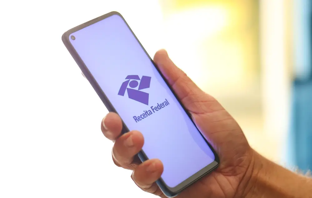 Mão segurando celular aberto na Receita Federal