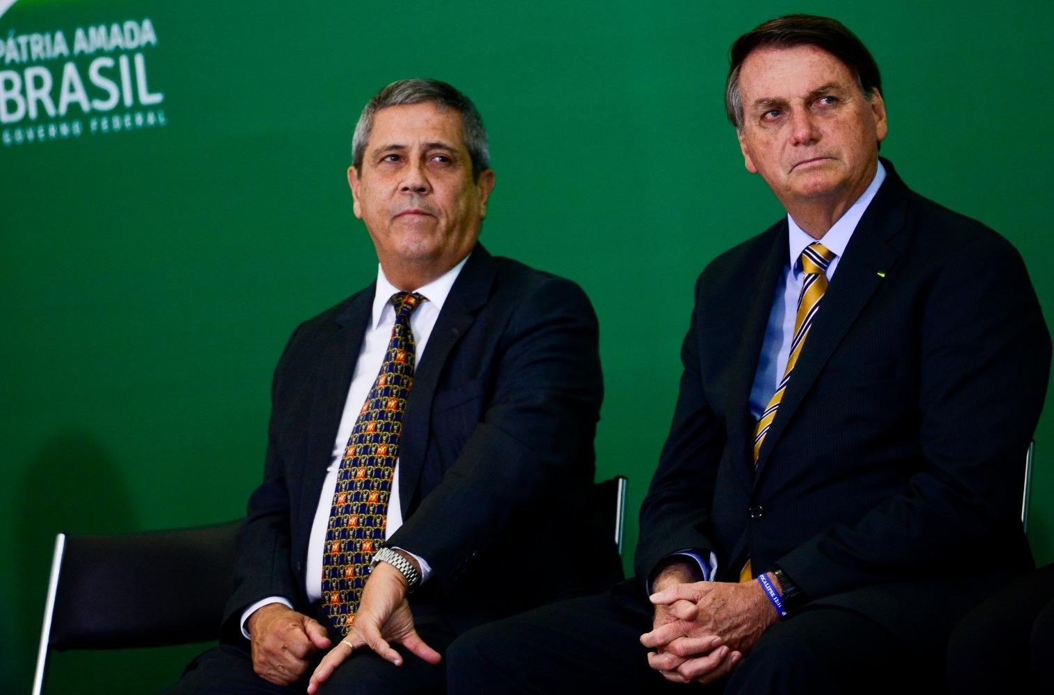 Braga Netto e Bolsonaro estão sérios observando alguém falar. Eles estão de terno e camisa social branca e azul