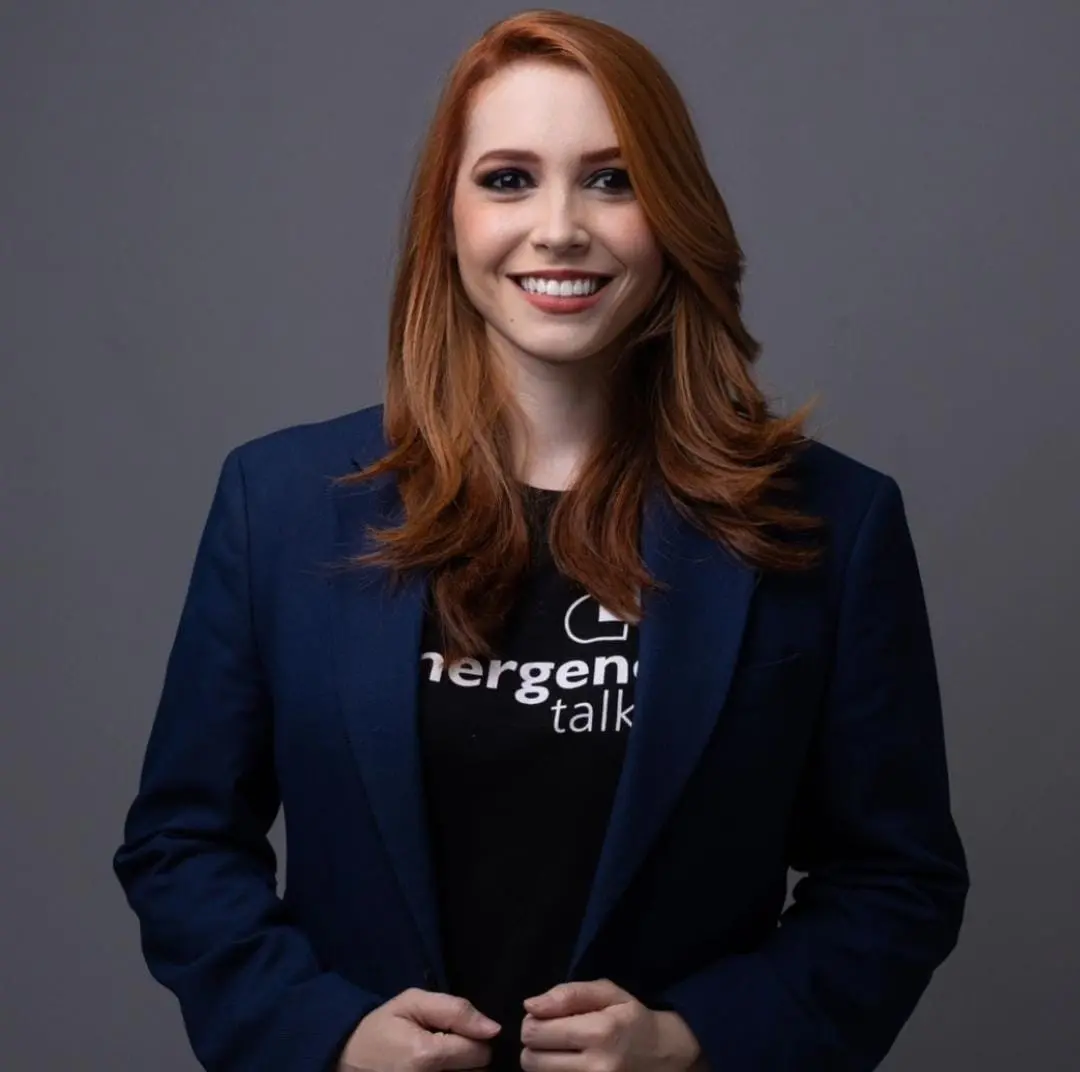 Patrícia é uma mulher branca de cabelo ruivo longo e liso. Na foto, ela está de camiseta preta e terno azul