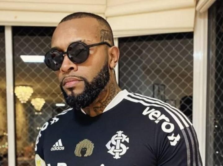 Nego Di é um homem negro de barba bem aparada com cavanhaque. Na foto, ele está com uma camisa preta d time e óculos escuros