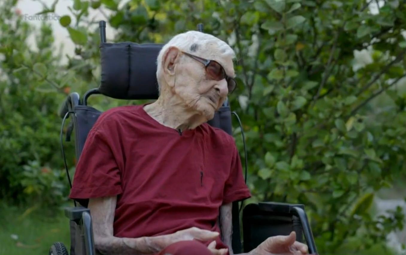 Foto do homem mais velho do mundo, o cearense João Marinho, de blusa vermelha, sentado em cadeira de rodas e de óculos escuros