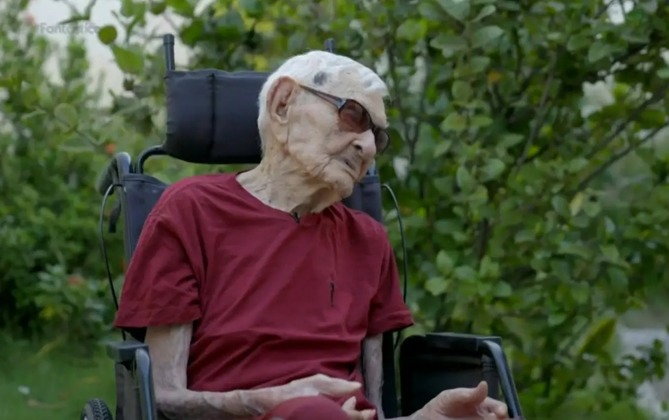Foto do homem mais velho do mundo, o cearense João Marinho, de blusa vermelha, sentado em cadeira de rodas e de óculos escuros