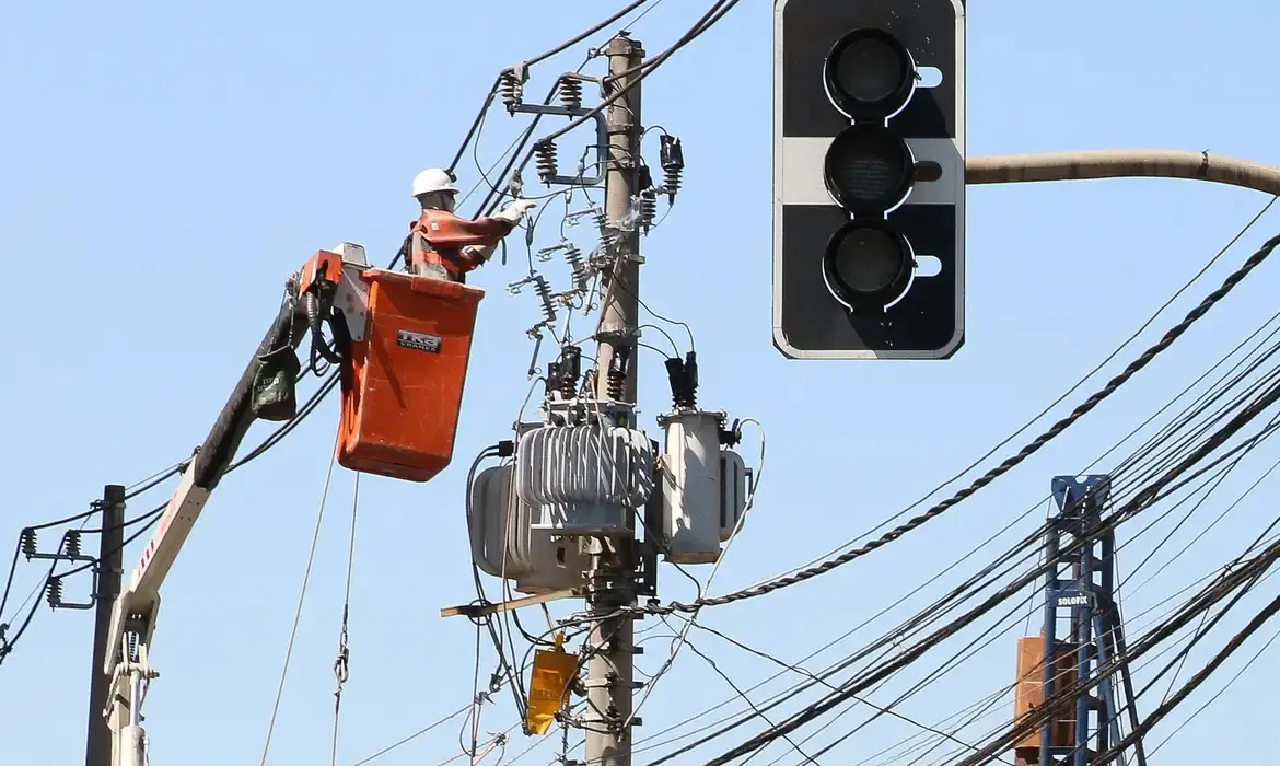 Homem trabalhando em altura em um caminhão de manutenção elétrica, ajustando fios e transformadores em um poste de energia sob um céu claro.