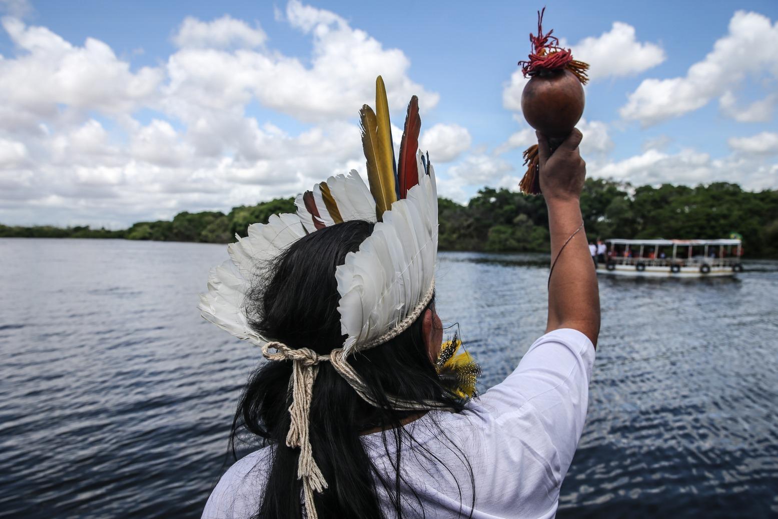 Indígena de costas com cocar e levantando chocalho