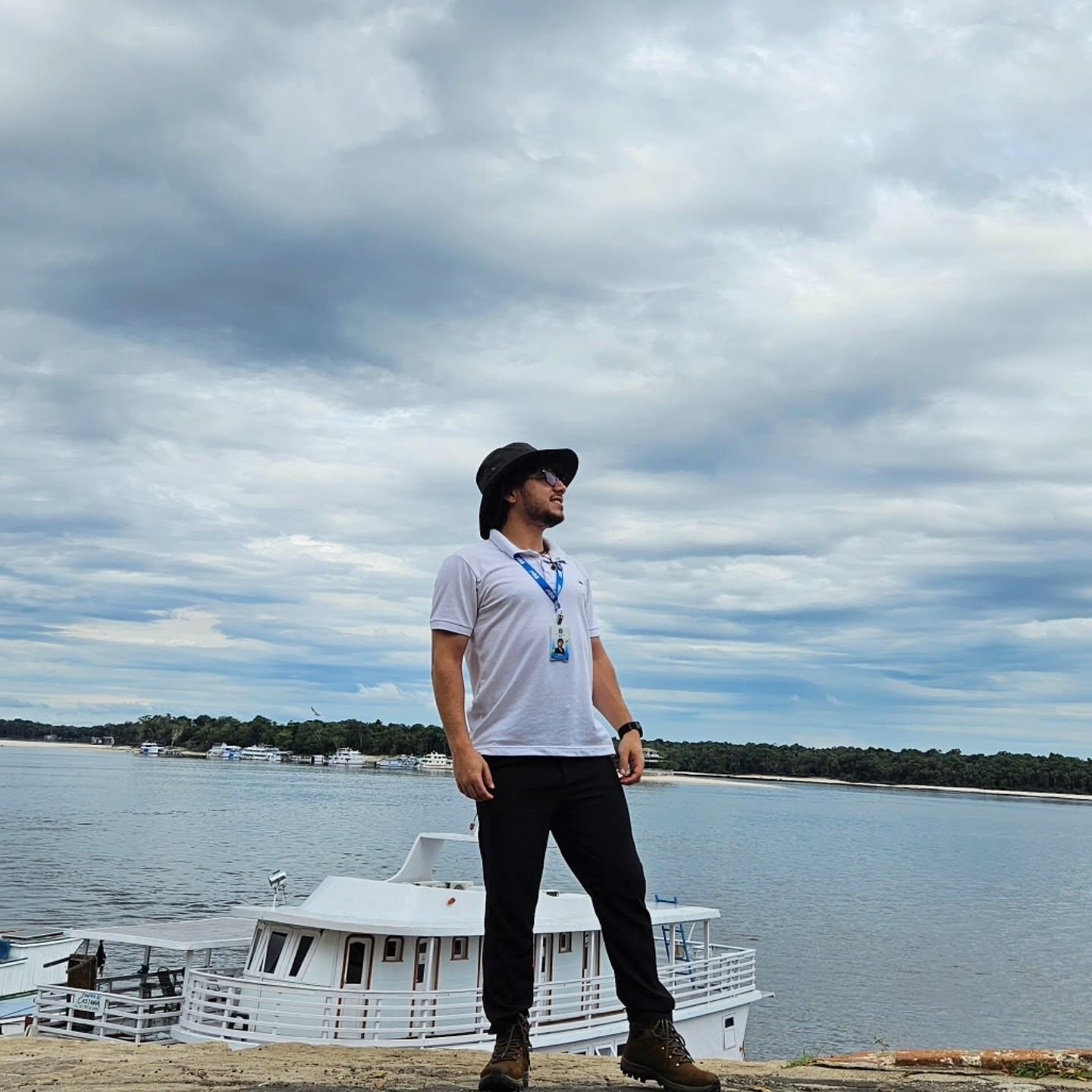 Homem em pé, olhando para o horizonte, com rio e barco branco ao fundo