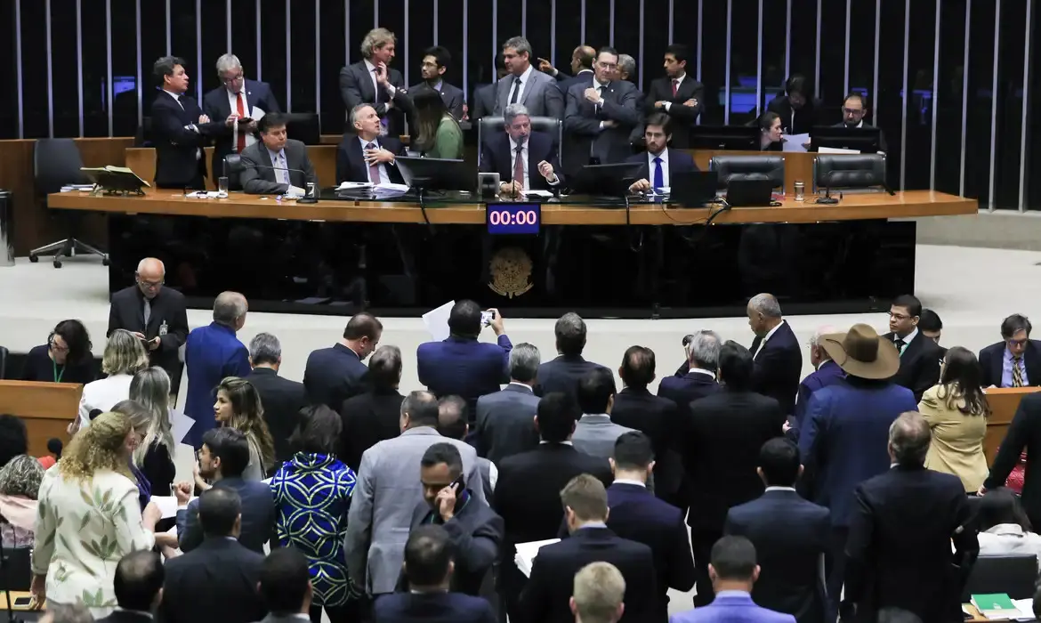 Imagem do plenário da Câmara dos Deputados durante uma sessão. Deputados conversam e debatem. O ambiente reflete a atividade parlamentar no Brasil.