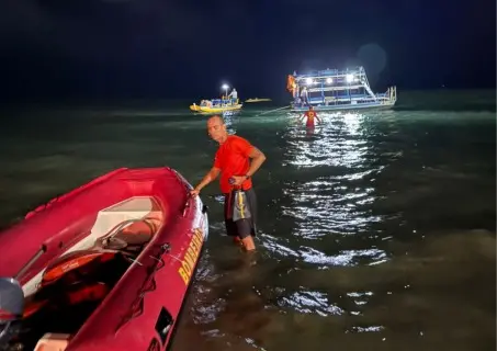 Imagem de um salva-vidas em ação durante a noite, ao lado de uma embarcação e um bote, no mar iluminado.