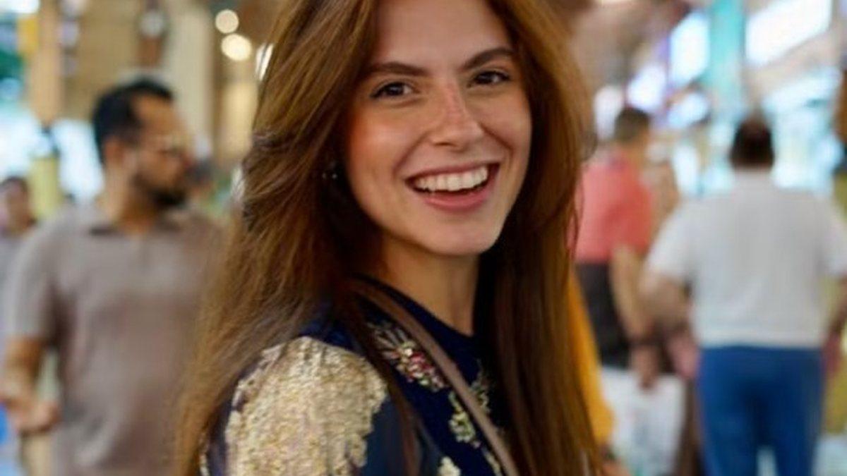 a imagem mostra jovem médica de Alagoas sorrindo com várias pessoas passando ao fundo da imaagem. Carolina faleceu neste domingo (29), aos 24 anos, em incêndio na Tailândia